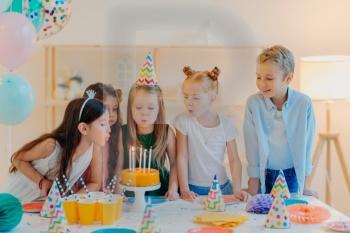 Small kids celebrate birthday party, blow candles on cake, gather at festive table, have good mood, enoy spending time together, make wish, wear party hats, pose indoor with inflated balloons