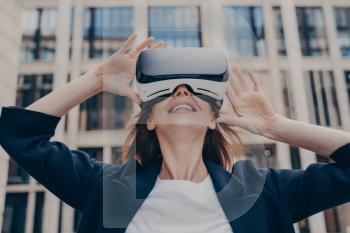 Immersed in virtual reality, futuristic redhead businesswoman wearing 3D goggles stands outside, looking up with awe, holding the portable VR glasses with both hands.