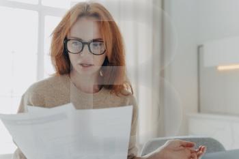 Serious redhead young woman focused in paper documents studies financial report wears spectacles casual jumper poses indoor against home interior. Busy ginger female financier does paperwork. Serious redhead young woman focused in paper documents studies financial report