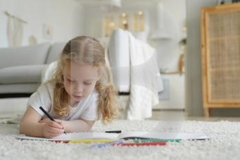 Talented little preschool child girl painting lying on warm floor in living room at home. Caucasian kid girl drawing with colored pencils playing spending leisure time alone. Children’s creativity. Talented preschool kid girl painting with colored pencils, lying on floor at home. Child creativity