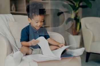 Happy mixed race little kid boy with cute curly hair reading story book while sitting in cozy armchair in light colored living room at home, child spending free time indoors. Children development. Happy mixed race little kid with cute curly hair reading book at home