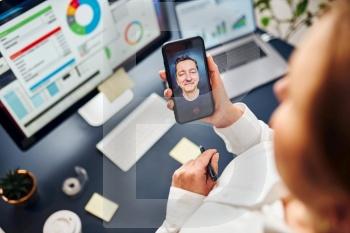Businesswoman having video chat on mobile phone with her colleague. Businesswoman working with data on charts, graphs and diagrams on computer screen. Woman holding smartphone sitting at desk in office