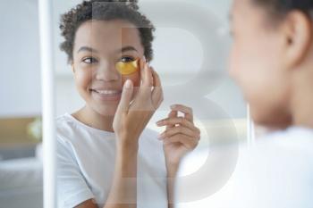Mirror reflection of young hispanic lady. Happy afro girl is applying eye patches to her face. Skin moisturizer with collagen serum. Anti age patches. Morning skincare. Beauty routine of teenage girl.. Mirror reflection of young hispanic lady with eye patches. Skin moisturizer with collagen serum.