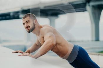 Bearded man does push-ups outdoors, warms up, poses near a bridge, has early morning workout.