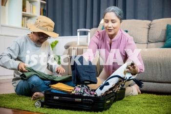 Asian romantic retired couple packing clothes travel bag suitcase together on floor at home interior living room, couple old senior married retired prepare luggage suitcase arranging for travel