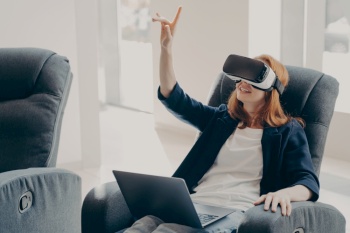 Young impressed redhead businesswoman using virtual reality glasses for business, female in VR headset pointing with finger in air, touching 3d object while working remotely on laptop from home. Female in VR headset pointing with finger, touching 3d object while working remotely on laptop