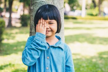The image of a little girl with her hands closed, her right eye leaning against a tree in a park.