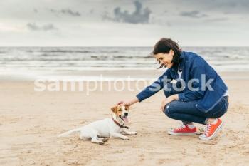 Brunette stylish woman teases her small dog on sea shore, have walk together, have real friendship, understand each other. Beautiful young female spends free time with pet outdoors. Dog and woman on seashore