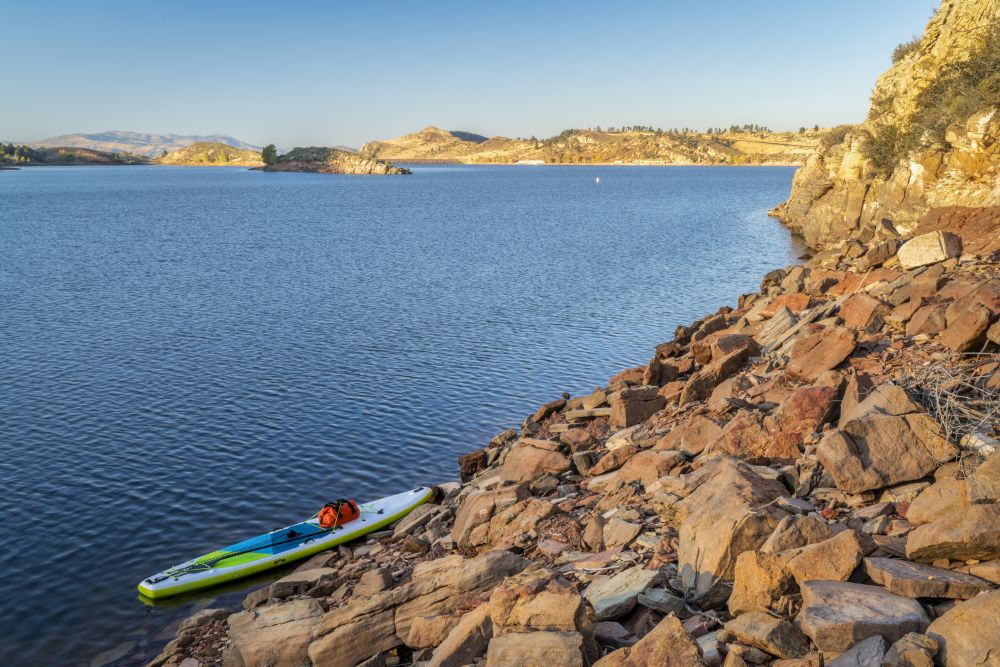 inflatable stand up paddleboard with a paddle and dry bag on a rocky shore of mountain lake - Horsetooth Reservoir in northern Colorado
