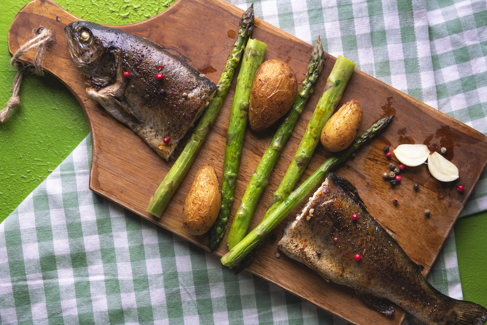 Baked trout, cut in half, seasoned with green asparagus and roasted potatoes, on a wooden cutting board and napkin, on a green background. Above view.