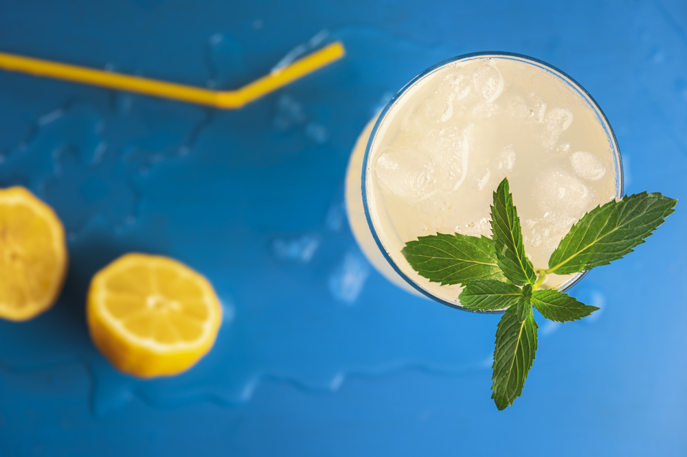 Above view of a lemonade glass with crushed ice and peppermint branch and yellow straw and lemons, on a blue background. Refreshing cold summer drink.