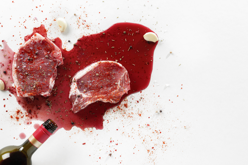 Cooking raw pork steaks with red wine and spices. Above view of wine bottle spilled over meat and seasonings on white background. Flat lay.