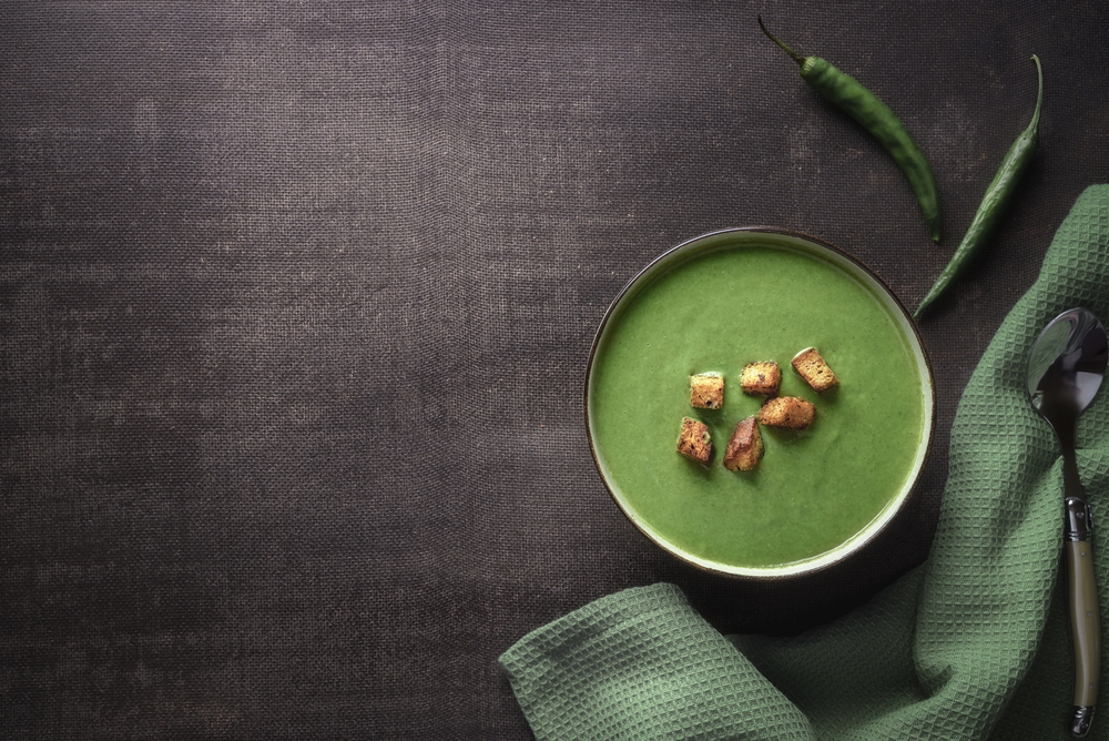 Cream soup in a bowl with spinach and green hot peppers and croutons, on a kitchen table. Above view of a vegetarian dinner table with spinach soup.