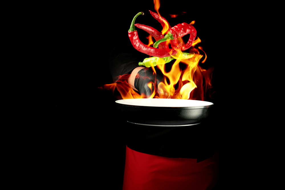 chef in a black uniform holds a round pan and throws up red and green whole chili peppers in a burning fire, cooking spices, low key, copy space
