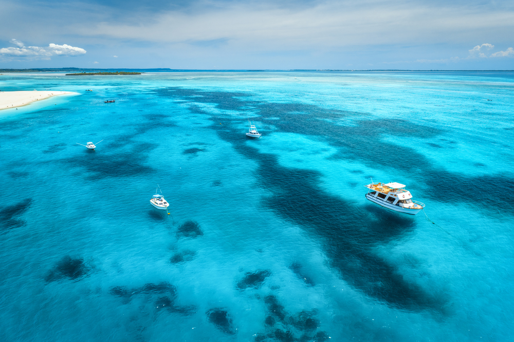 Aerial view of the yachts, fishing boats in transparent blue water at sunny day in summer. Top view of boat, sandy beach, sky. Travel in Zanzibar, Africa. Colorful landscape with motorboats in the sea