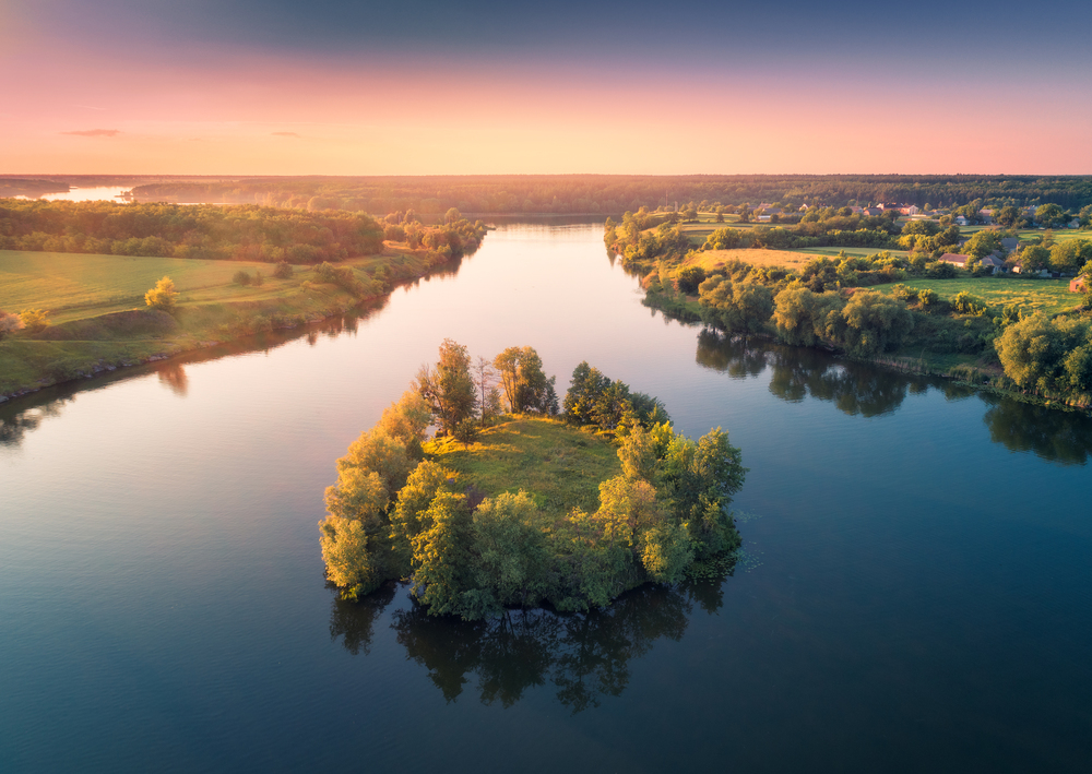 Aerial view of beautiful small island with green trees in the river at sunset in summer. Colorful landscape with island, meadow, forest and sky reflection in blue water. Top view from air. Nature  . Aerial view of small island with green trees in the river