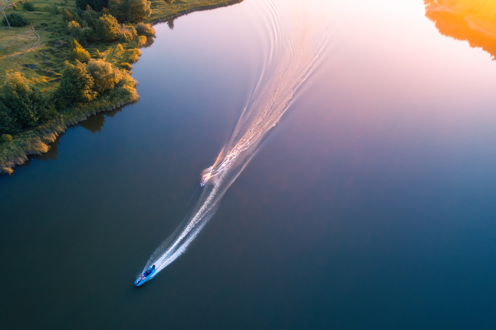 Aerial view of floating water scooter in blue water at sunset in summer. Holiday in countryside. Top view of boats in motion in the river. Landscape with moving motorboat and green trees. Extreme. Aerial view of floating water scooter in blue water at sunset