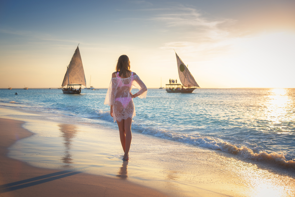 Beautiful young woman is standing in sea with waves on sandy beach against sailboats at sunset. Summer travel. Tropical landscape with slim girl in white lace dress on the seashore, boats and yachts