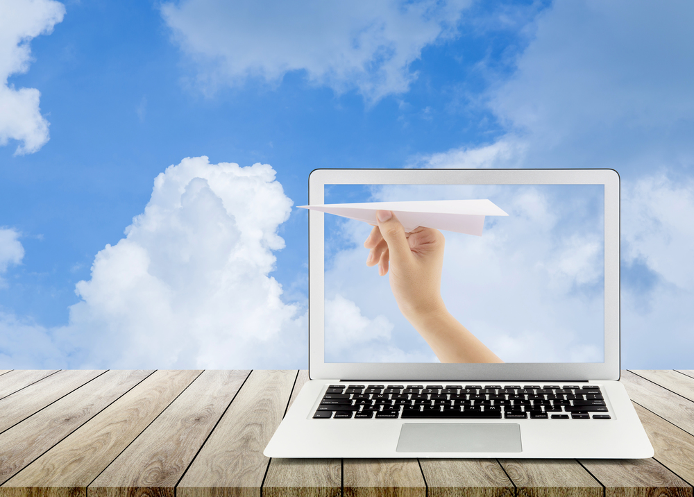 Hand with paper plane with laptop on wood table against blue sky sending email, communication concept.