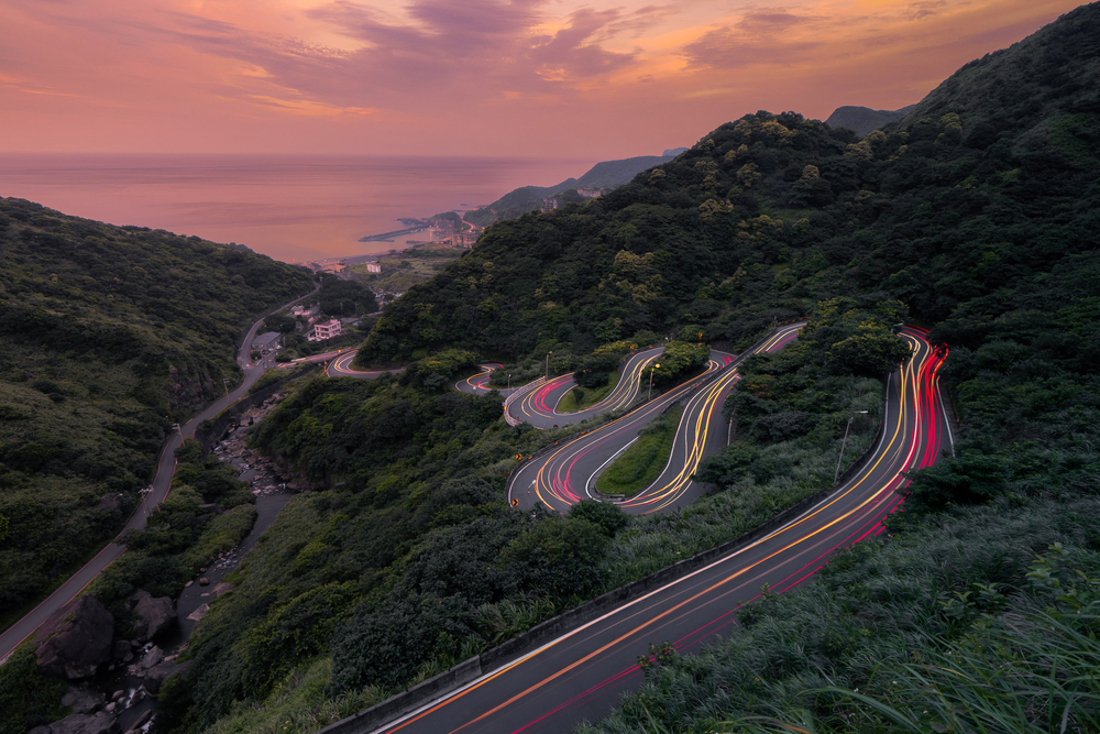 Aerial view of cars driving on curved, zigzag road or street on mountain hill with natural forest trees in rural area of New Taipei City, Taiwan at sunset