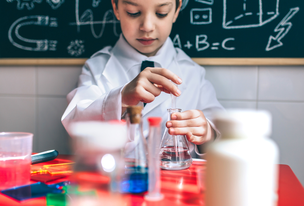 Portrait of little boy scientist extracting liquid from flask against of chalkboard with drawings. Selective focus on flask.. Serious kid playing with chemical liquids