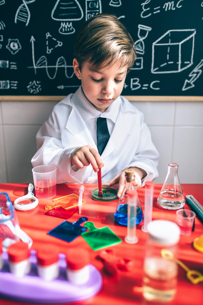 Portrait of little boy scientist mixing colorful chemical liquids against of chalkboard with drawings. Kid playing to be chemist with colorful liquids