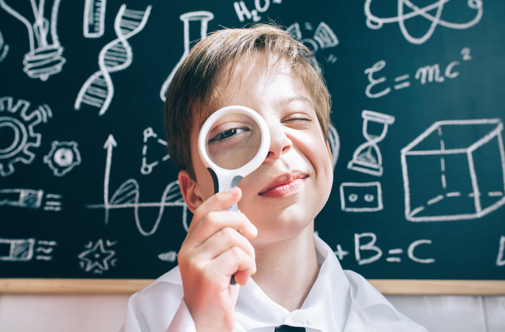 Close up of curious little scientist looking through magnifier against of drawn blackboard. Little boy looking at camera through magnifying glass