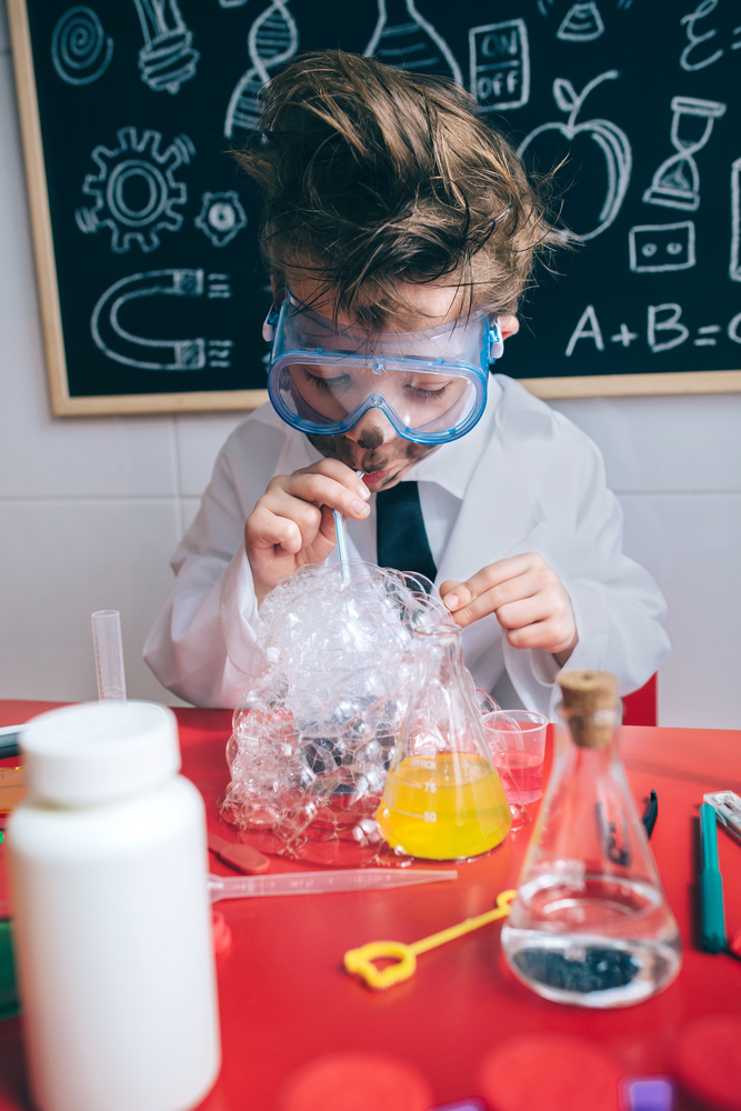 Crazy little boy scientist with glasses and dirty face doing soap bubbles with straw into glass. Kid doing soap bubbles with straw in glass