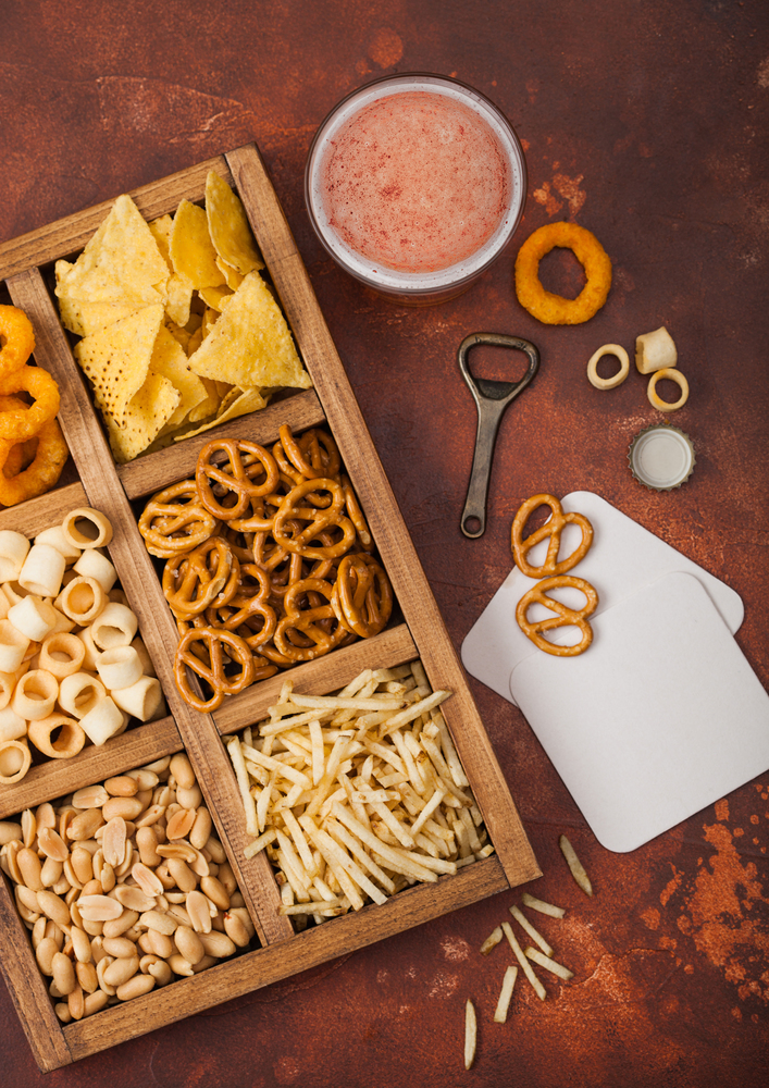 Glass of craft lager beer and opener with box of snacks on brown kitchen background. Pretzel,salty potato sticks, peanuts, onion rings with nachos in vintage box with openers and beer mats.