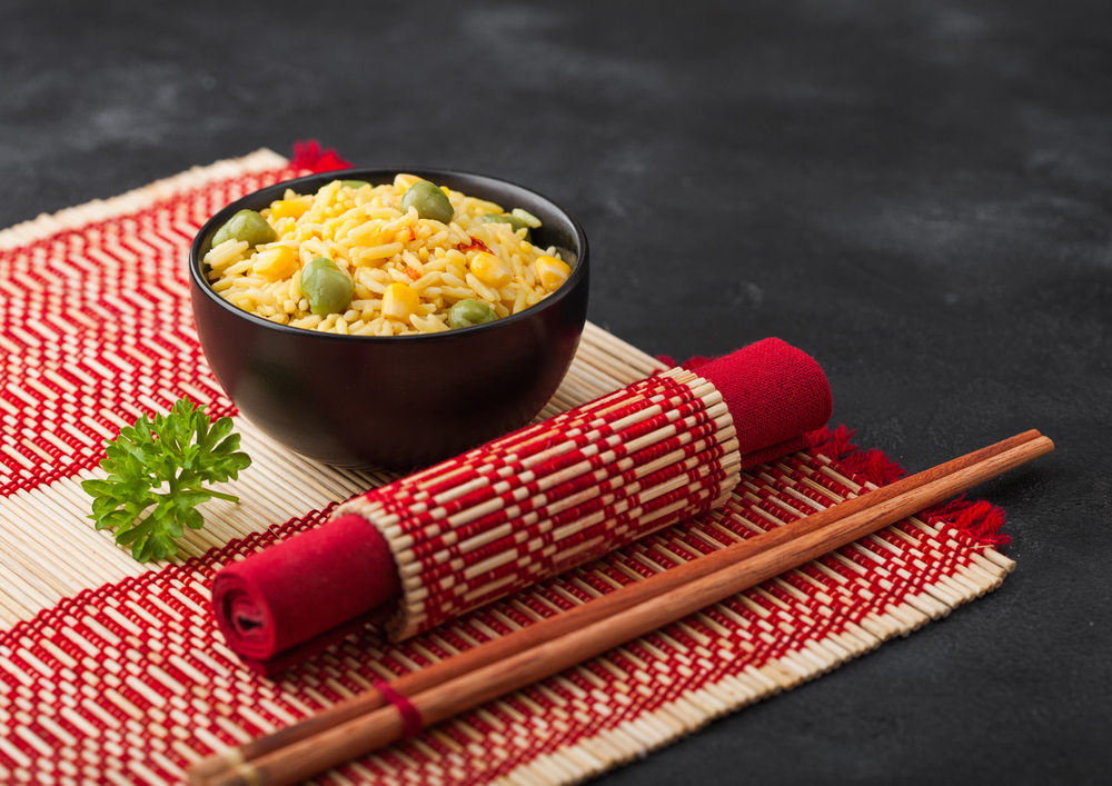 Black bowl with boiled organic basmati vegetable rice with wooden chopsticks on red bamboo placemat. Yellow corn and green peas with paprika.