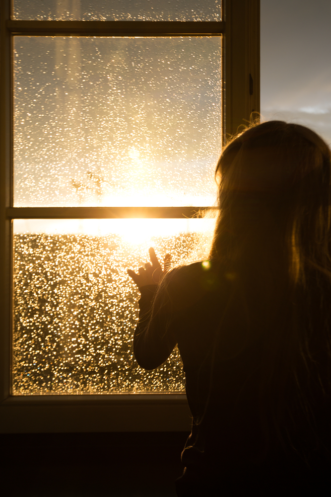 girl sitting on a windowsill and looking at the evening city at the beams of orange sunset sun