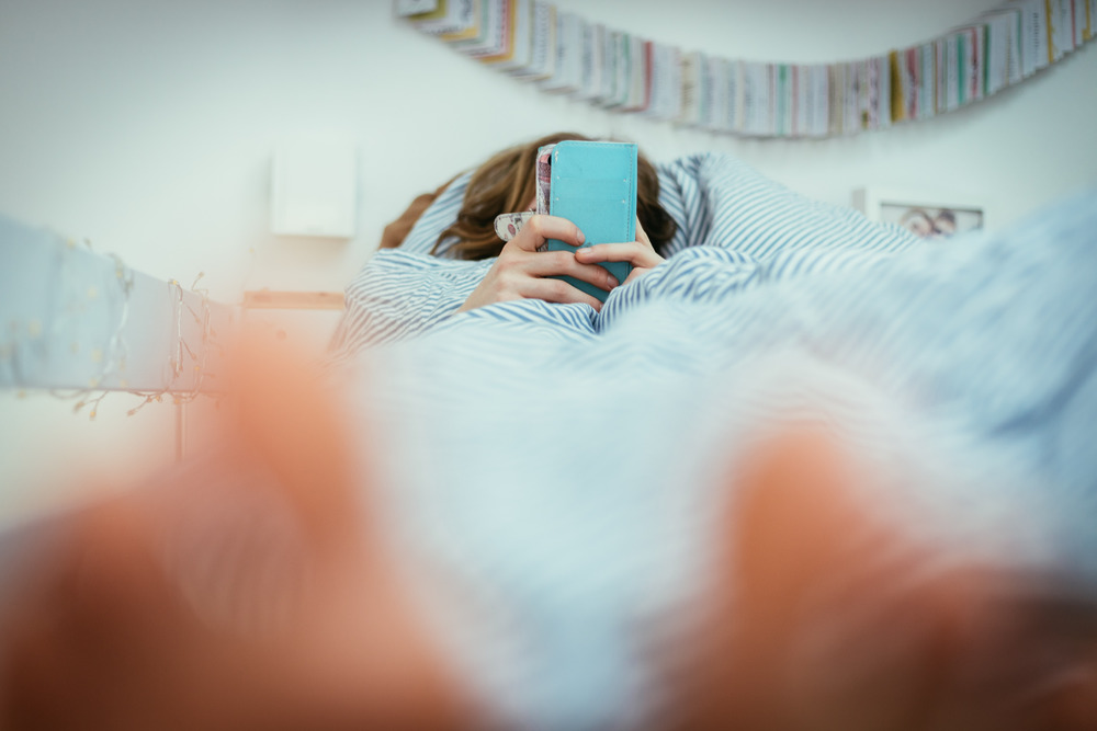 Girl lying in bed is using her smartphone, smartphone in the foreground, blurry background