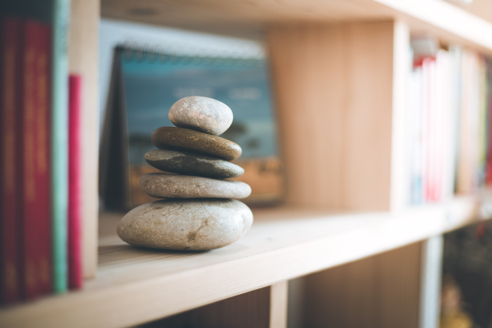 Feng Shui: Stone cairn at home in a book shelf, blurry books in foreground and background. Balance and relaxation. Sunlight.