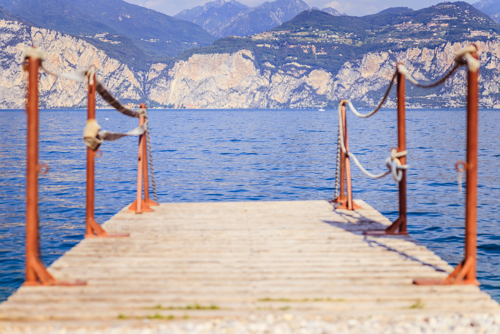 Wooden dock pier extending over blue lake water, mountains at lago di garda. Text space.