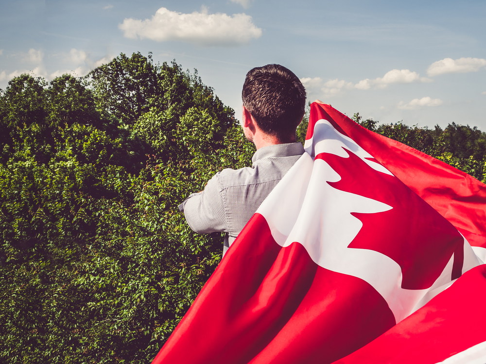 Handsome man waving a Canadian Flag against a background of trees and blue sky. View from the back, close-up. National holiday concept. Man waving a Canadian Flag. National holiday