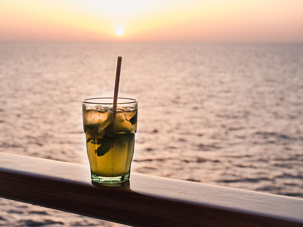 Beautiful glass, bright cocktail with ice and straw, standing on the open deck against the backdrop of sea waves and sunset. Side view, close-up. Concept of leisure and travel. Beautiful glass and bright cocktail with ice