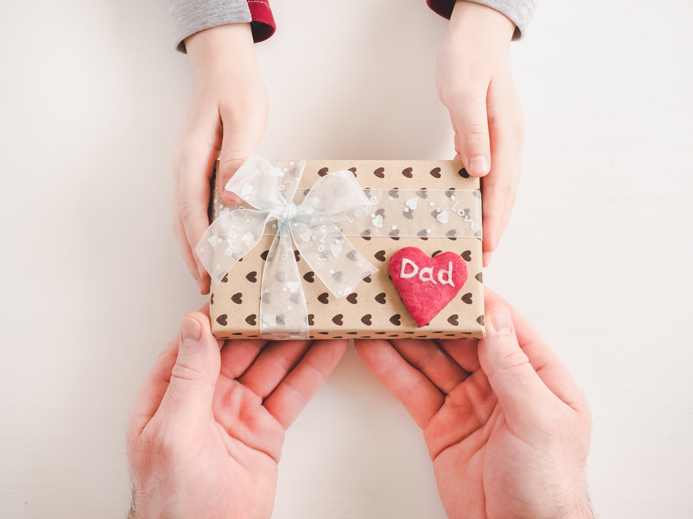 Child&rsquo;s hands and adult man&rsquo;s hands, beautiful gift box, ribbon and glazed cookies on a white, wooden background. Top view, close-up. Preparing for the holidays. Beautiful card for Women&rsquo;s Equality Day. Close-up