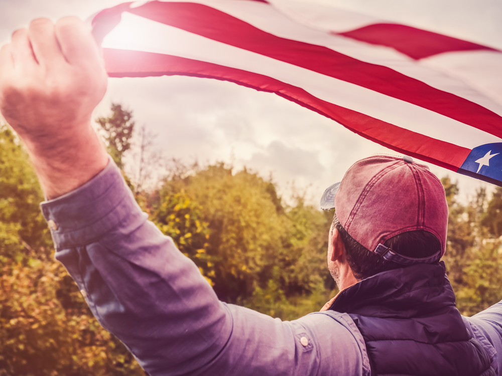Handsome man waving an American flag against a background of trees and blue sky. View from the back, close-up. National holiday concept. Handsome, young man waving an American flag