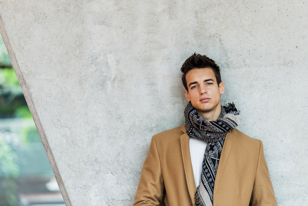 Front view of stylish young man wearing coat and scarf leaning on a wall while looking camera