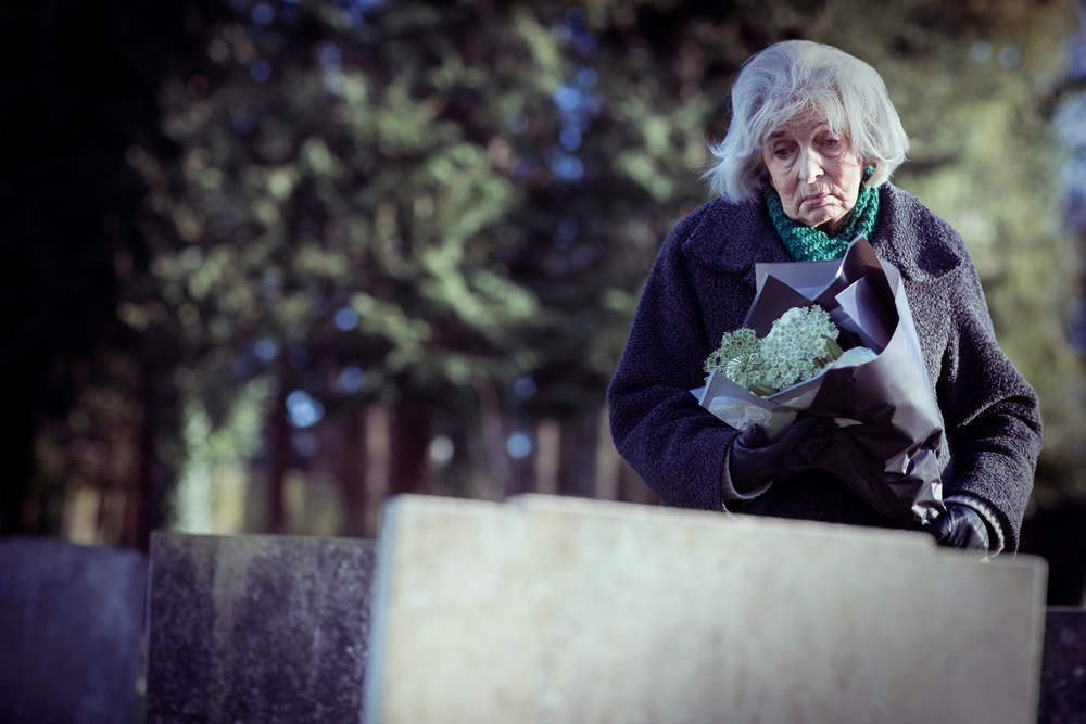Sad Senior Woman With Flowers Standing By Grave