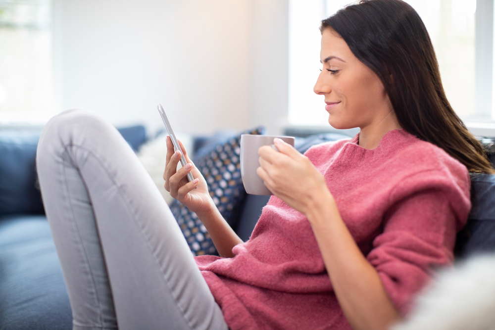 Young Woman Relaxing On Sofa With Hot Drink Checking Mobile Phone