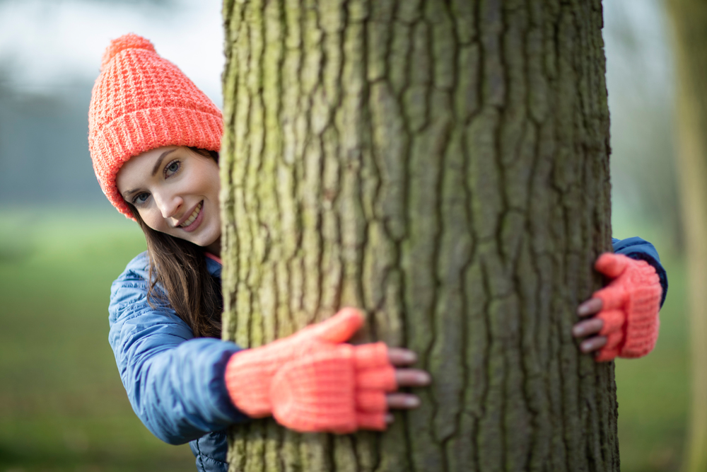 Portrait Of Woman Wearing Gloves Hugging Tree Trunk On Winter Walk In Countryside