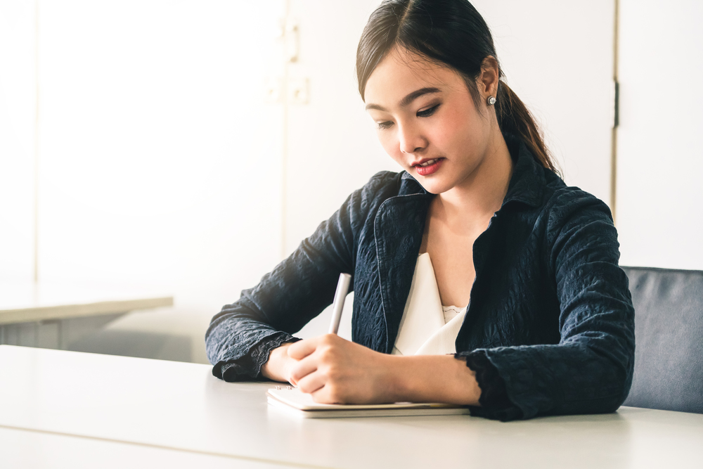 Beautiful young Asian woman writes letter on notebook while sitting at office desk. Content writer and secretary job concept.