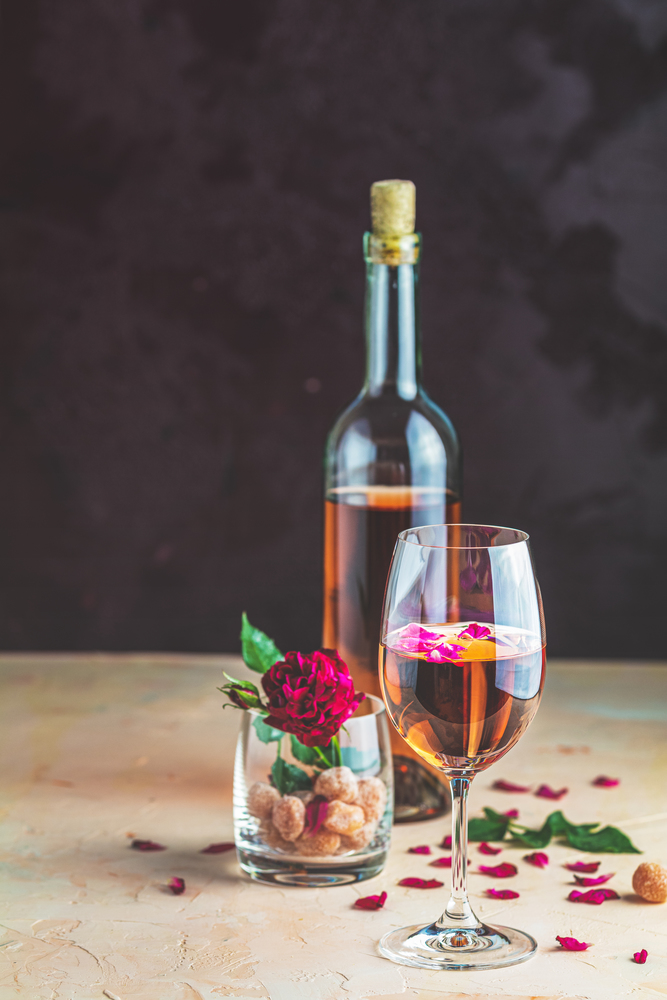 Bottle of rose wine and glass served with rose wine and rose petals, rose and candied fruit in glass on pink concrete table and dark background. Beautiful valentine or wedding greeting card.