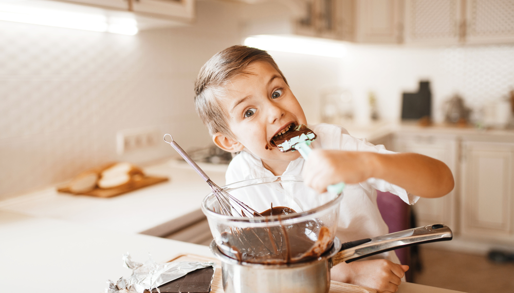 Young boy tastes melted chocolate in a bowl. Kid cooking on the kitchen. Happy child prepares sweet dessert at the counter