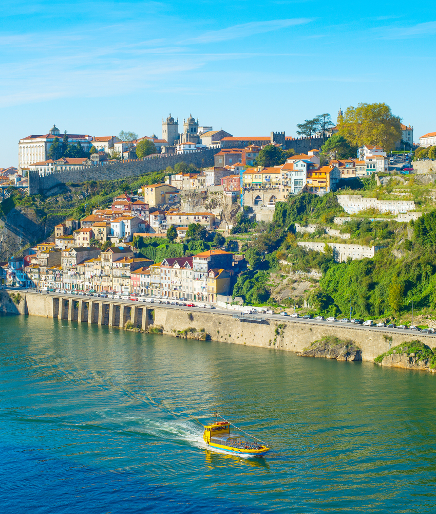 Tourist boat sailing by Douro with Porto cityscape on green riverbank in bright sunlight, Portugal