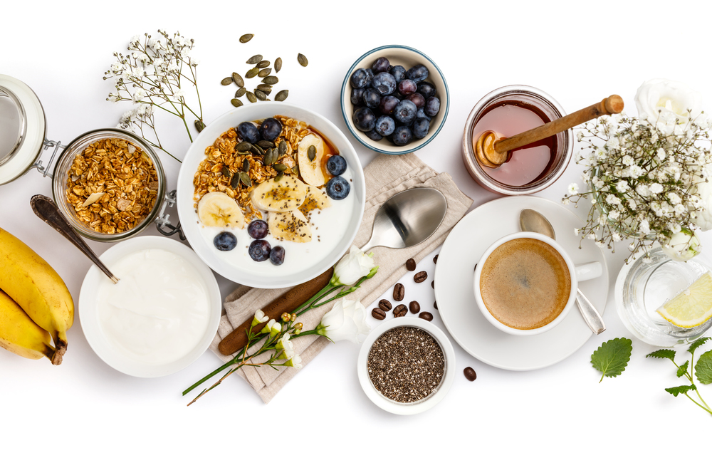 Healthy breakfast set. Oat granola with yogurt, honey, fresh bananas, blueberries, chia seeds in bowl  and cup of coffee on white background, top view. Healthy breakfast set on white background, top view, copy space