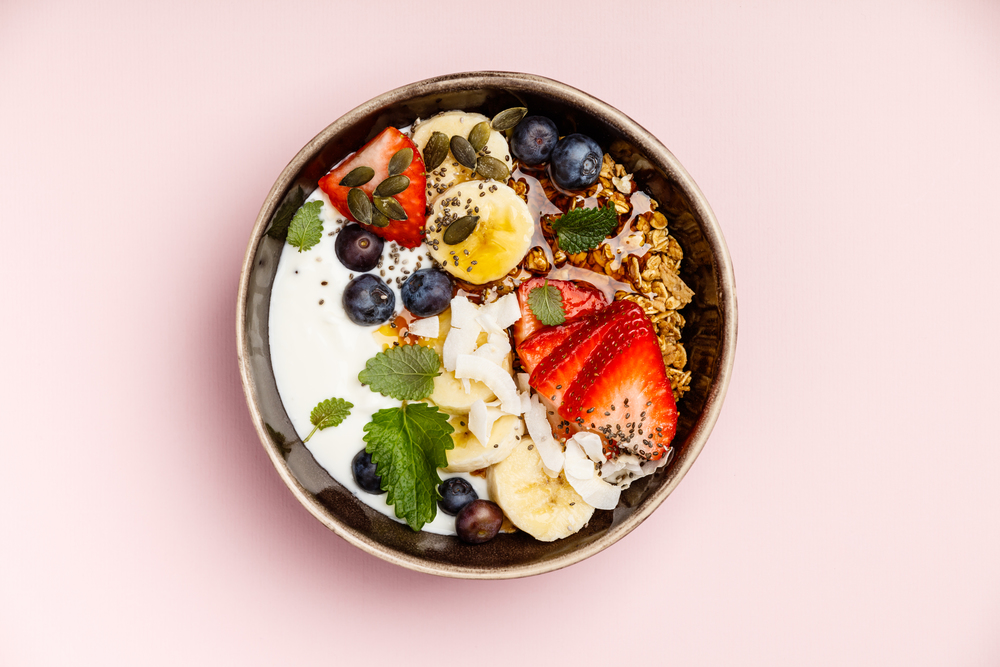 Healthy vegetarian breakfast- Oat granola with fresh berries, banana, yogurt, maple syrup, seeds and mint leaves on pink background, flat lay, top view. Healthy breakfast set on pink background, top view, copy space
