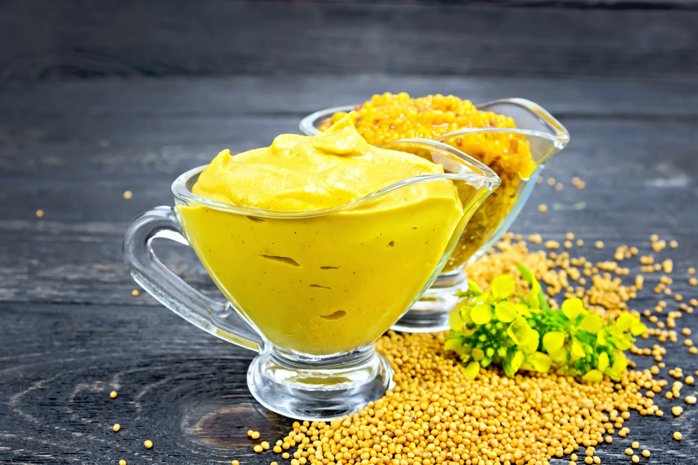 Mustard sauce and Dijon mustard in two glass saucepans, yellow flower and seeds against a black wooden board. Sauce mustard in two sauceboats with flower on table
