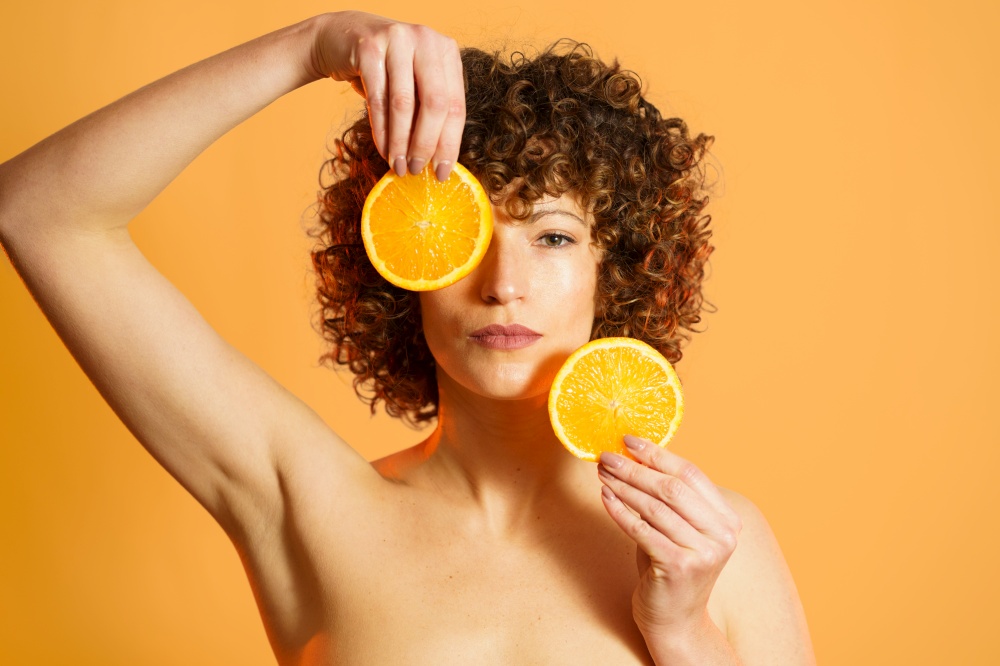 Serious adult female with curly hair covering face with slices of ripe citrus during beauty routine against orange background. Adult woman with pieces of fresh orange